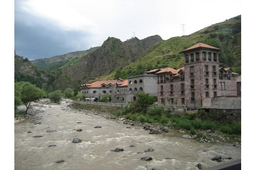 A castle-like building sits beside a rushing river and in front of green mountains.