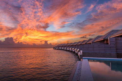 Standing on the edge of a floating pool at sunset, showing the perspective of multiple villas with their own floating pools stretching out before you. Sunset of bright orange, yellow, and purple.