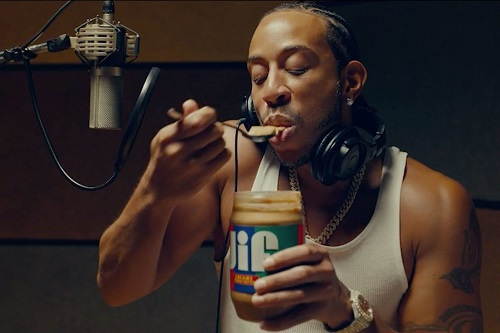 Rap artist Ludacris takes a spoonful of Jif peanut butter from the jar while standing in front of a microphone in a recording studio.