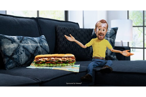 A picture of a subway foot-long sandwich and the mascot from Bob's Discount Furniture sitting on a blue sectional sofa.