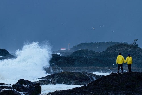Two people in yellow rain gear stand at the edge of the ocean with rough seas as part of the Wickaninnish Inn's Storm Watching package, which is an excellent example of how to promote your brand's weaknesses strategically.