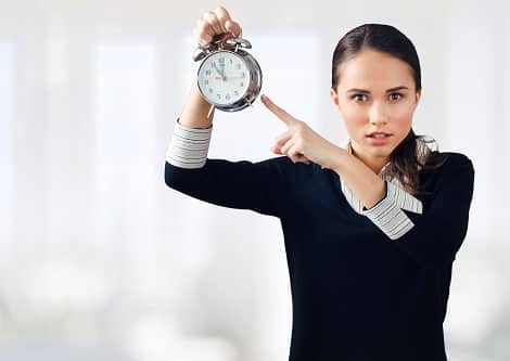 A woman wearing a black sweater with grey cuffs and collar holds up a clock and points to it, implying that time pressure is one factor in how to stop being an impatient writer.