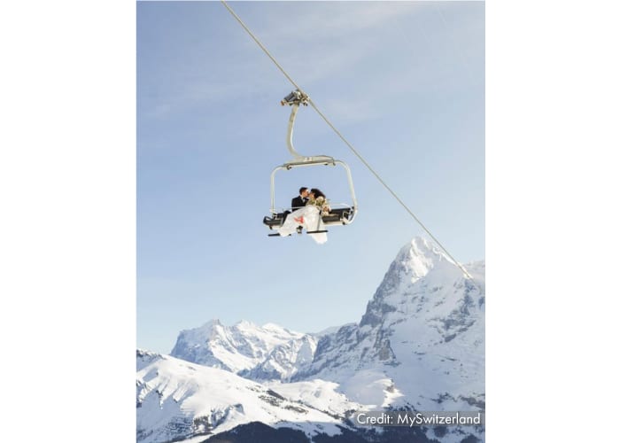 A bride and groom dressed for a wedding sit high up on a ski lift chair with the white snowy Alps in the background, as an example of what makes a dramatic tourism marketing photo.