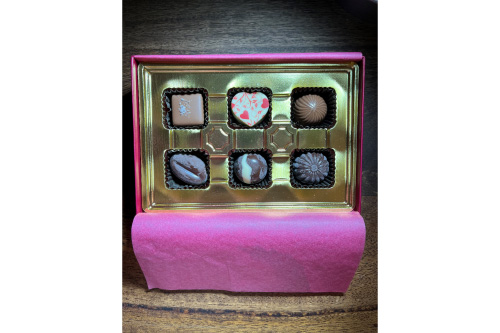 A gold box with pink tissue paper houses six individual chocolates, two milk, three dark, and one white one shaped like a heart with red designs on it.