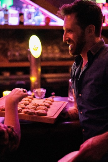 A man with a blue shirt holds a wooden board with chocolate chip cookies on it, serving it to a customer whose arm can be seen picking a cookie off the board. This is at Too Soon, a cocktail bar in Portland, Oregon, where they serve these cookies every night before last call. It's an excellent example of how small touches can make a big impact in hospitality.