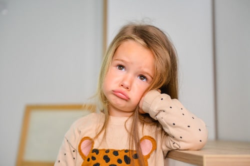A girl toddler with long blond hair wearing a beige long sleeved shirt rests her head on her hand and wears a pouty, sad expression. 