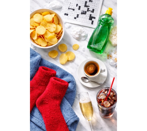 An aerial view of several items that could help someone become an idea person, including a bowl of potato chips, red fuzzy socks, green dishwashing liquid, a cup of espresso, a glass of champagne, a glass of coke, and a crossword puzzle.